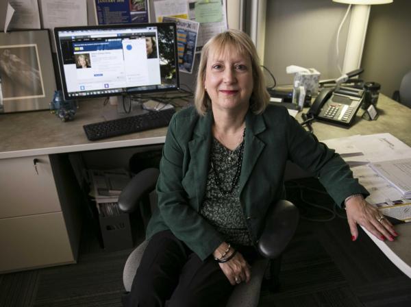 The image shows a woman sitting in an office chair at her desk, smiling at the camera. She is wearing a green blazer and black pants. Her desk is organized, with a computer monitor displaying a professional profile. The workspace includes a phone, papers, and personal items, creating a professional yet personalized environment. The background has various documents and a framed photo, adding to the office's detailed and lived-in feel.