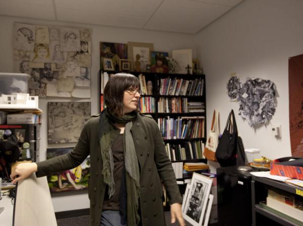 The image shows a woman standing in a cluttered office or studio space. She is wearing a dark coat and a scarf, with glasses and shoulder-length dark hair. The room is filled with bookshelves, art supplies, and various pieces of artwork, including sketches on the walls and framed art on shelves. The woman appears to be in the midst of moving or organizing, as she is looking to the side with her hand resting on a chair. The space has a creative and personal atmosphere.