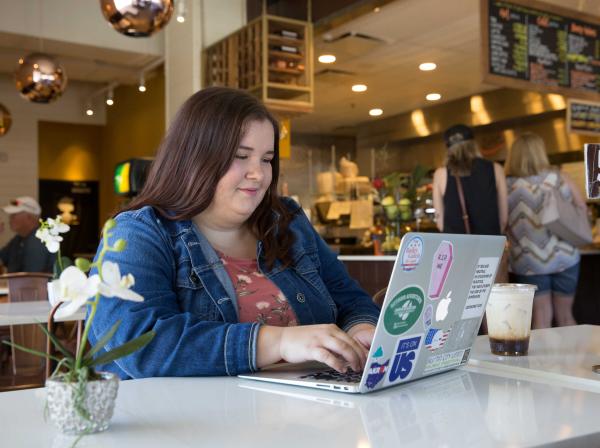 A student types on their laptop and sits at a table in a cafe.