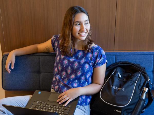 A student sits on a bench with a laptop in their lap and backpack by their side and looks out the window.