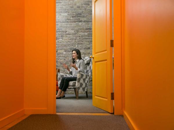 The image shows a woman sitting in an armchair in a room with bright orange walls and an open door. The photo is taken from the hallway, looking into the room through the doorway. The woman is engaged in a conversation, gesturing with her hands, and is wearing a gray sweater and black pants. The room has a brick wall, adding a rustic feel to the modern, brightly colored space. The scene captures a candid moment in what appears to be an informal, cozy setting.