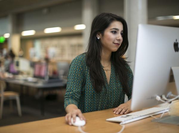 The image shows a woman sitting at a desk, using a large Apple desktop computer. She is holding a computer mouse and looking at the screen with a focused expression. The background appears to be a modern, open-plan office or study space, with other people working at desks in the distance. The woman is wearing a green patterned top, and the environment suggests she might be in a library or co-working space.