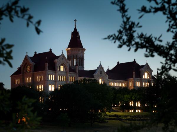 Main Building is framed by foliage in the foreground with windows illuminated at dusk.