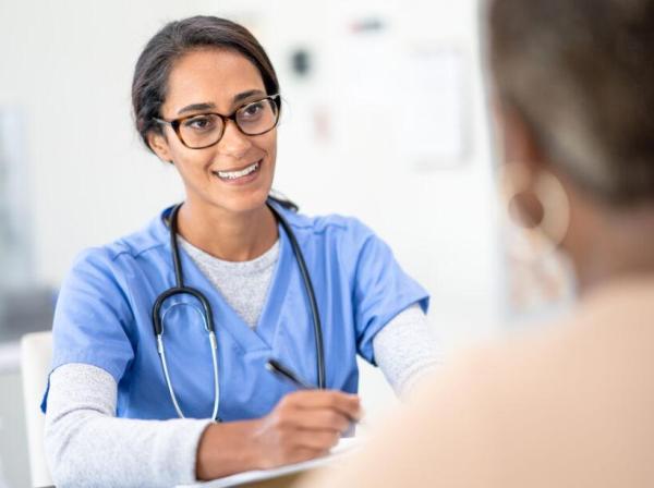 The image shows a healthcare professional, likely a nurse or doctor, engaged in a conversation with a patient. The medical professional is wearing blue scrubs, glasses, and a stethoscope around her neck, and she is smiling warmly, holding a pen and possibly taking notes. The setting appears to be a clinical environment with a bright, clean background. The patient's shoulder is visible in the foreground, with the focus on the healthcare professional.