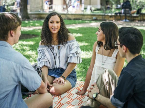 The image shows a group of four young adults sitting on a blanket on a grassy area, likely in a park or on a campus. They are engaged in a casual conversation. The group includes two women and two men. One of the women, with curly hair and wearing an off-the-shoulder top, is smiling as she speaks. The setting is bright and outdoors, with trees and other people in the background, suggesting a relaxed and social environment.