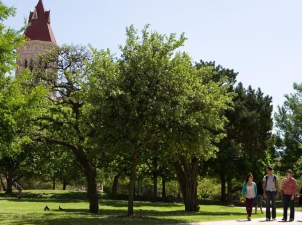 5 students are walking at the St. Edward's Campus with a background that includes trees and the Main Buidling.