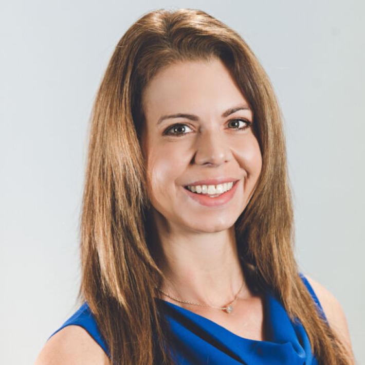 The image shows a woman with long, straight light brown hair wearing a sleeveless blue dress with a cowl neckline. She has a friendly expression with a broad smile and is facing slightly to her right. The background is a plain light gray. She has light skin and an approachable demeanor.