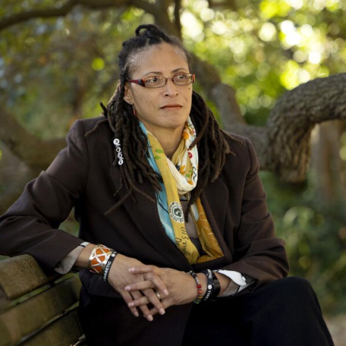 A woman wearing glasses, bracelets, a jacket, and a scarf sits on a bench under a tree