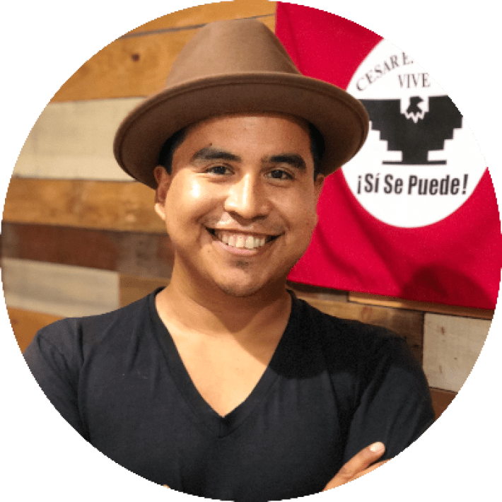 Close-up headshot of a man wearing a hat, standing in front of a flag that says "si se puede"