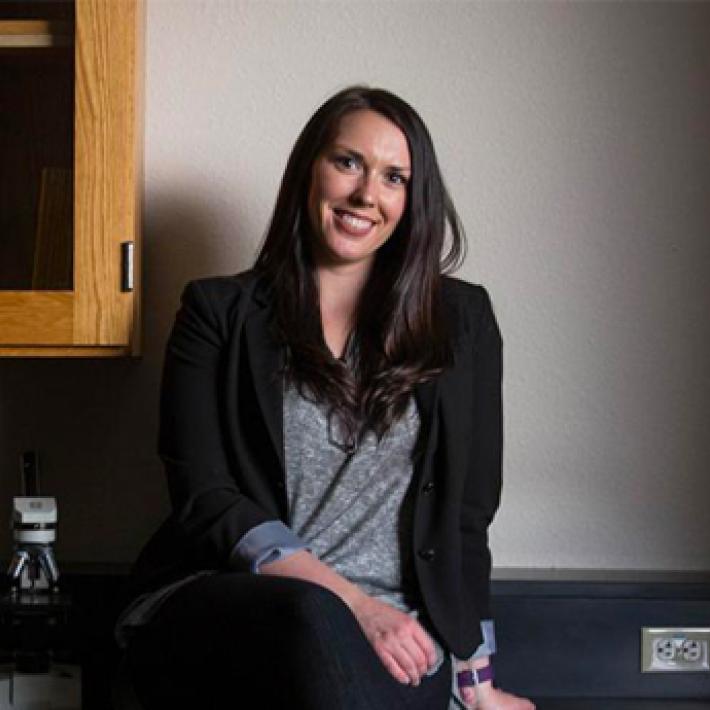 The image shows a woman with long, dark hair sitting on a counter in a room that appears to be a laboratory or classroom. She is wearing a black blazer over a patterned blouse and black pants. She has a friendly expression with a broad smile and is looking directly at the camera. Behind her are wooden cabinets and a small section of a tiled wall. The lighting is soft, creating a warm and approachable atmosphere.