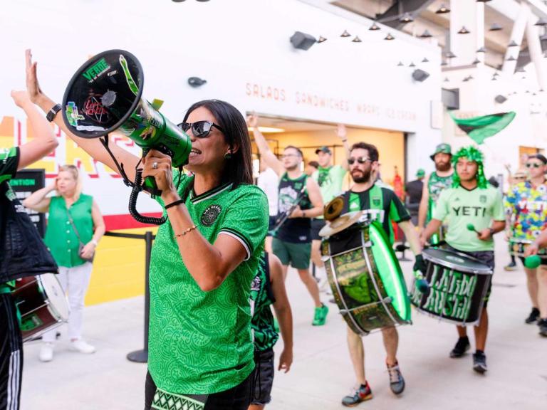 The image shows a group of enthusiastic fans, wearing green and black attire with "YETI" logos, celebrating and marching with drums and a megaphone inside a stadium. The scene is lively, with one person leading chants through the megaphone while others play drums and high-five. The overall atmosphere is energetic and festive, indicating a sports event or rally.