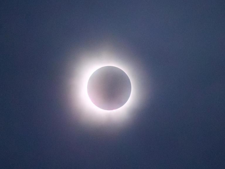 The image captures a solar eclipse in its totality phase, where the moon completely covers the sun, leaving only the bright corona visible as a glowing ring around the darkened disk. The sky appears dark, adding to the dramatic effect of the eclipse. The soft, glowing light of the corona creates a striking contrast against the shadowed moon, making this a captivating depiction of a natural celestial event.