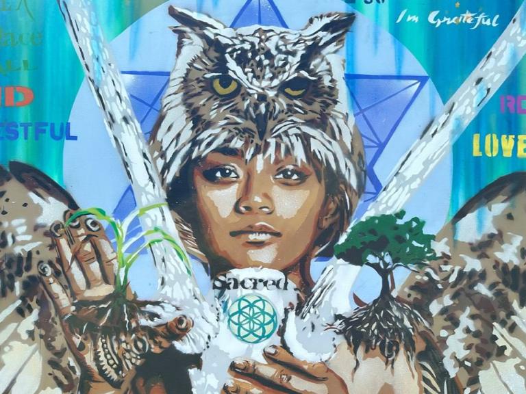 The image is a vibrant mural featuring a young person's face with an owl perched on their head. Their hands hold various elements: a plant, a tree, and a white object with the word "sacred" and a geometric design. The mural incorporates words like "kind," "restful," "love," "respectful," and "beautiful," along with phrases like "you are" and "I'm grateful." The background features a blue and green gradient with geometric shapes, creating a theme of nature, wisdom, and positive affirmations.