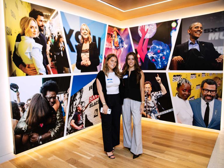 Students pose in front of a wall of photos from SXSW while visiting their offices in Austin for internships.