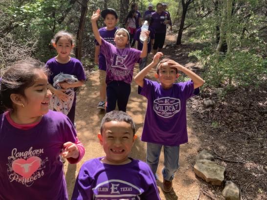 The image shows a group of children on a nature trail. They wear t-shirts with various prints, including "Longhorns Princess" and "Wildcats." Trees and foliage surround the trail.