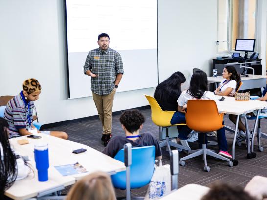 A staff member stands at the front of a classroom and speaks to students seated at tables during orientation.