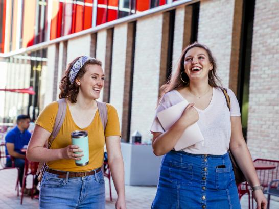 Two young women walking and laughing outdoors in an urban setting with brick buildings and glass windows in the background. One woman has curly hair and is wearing a mustard yellow shirt with jeans and a headband, carrying a backpack and holding a tumbler. The other woman has long hair, is wearing a white shirt with a denim skirt, and is carrying a folder or laptop. Both appear to be students, enjoying a casual and cheerful moment.