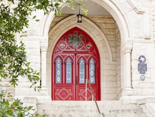 The red doors of Main Building, adorned with intricate stained glass windows and ornate designs. The doors are set within a stone archway. There are stone steps leading up to the entrance, flanked by columns and foliage partially framing the scene. Plaques are mounted on the stone wall beside the doors, indicating historical or informational significance. The overall atmosphere is one of elegance and tradition.