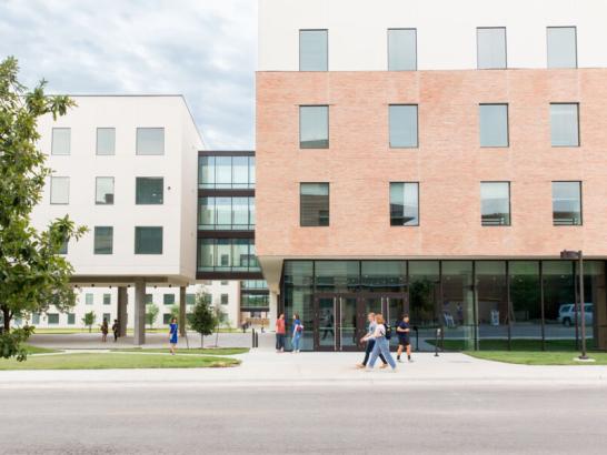 The image shows a modern multi-story building with a brick and light-colored exterior. Large windows line the building, and the ground floor features expansive glass panels. People are walking on the sidewalk in front of the building, contributing to a lively atmosphere. The building complex includes a connecting structure with additional windows, creating a cohesive design. The surroundings are well-maintained, with trees and grassy areas visible. The setting appears to be part of a campus.