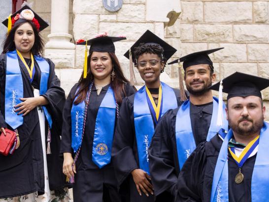 Graduates wear caps and gowns and veteran stoles and stand on the steps of Main Building.