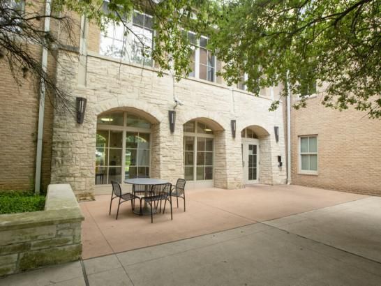 The image shows an outdoor courtyard area of a building with a stone and brick exterior. The courtyard features a small seating area with a round table and four chairs. The building has large windows and arched doorways, creating a welcoming and open feel. The space is shaded by overhanging tree branches. The courtyard is paved, with a stone bench on one side, and appears to be a quiet, inviting spot for relaxation or socializing and ideal for outdoor gatherings or a peaceful retreat.