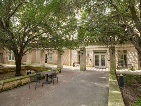 This image depicts an outdoor courtyard area surrounded by brick buildings with stone accents. The courtyard features a walkway leading to a building entrance with double doors. There are trees providing shade, and a small seating area with metal tables and chairs is positioned near the walkway. The scene appears serene and inviting, suggesting a quiet spot for relaxation or studying within a campus or residential complex.