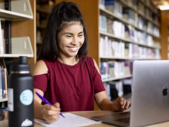 The image shows a young woman with long, dark hair tied up in a high ponytail, sitting in a library. She is wearing a red shirt with cut-out shoulders and is smiling while working on a laptop. She has a pen in her hand and is writing on a piece of paper. There is a large water bottle on the table beside her. Bookshelves filled with books are visible in the background, indicating she is in a study environment.