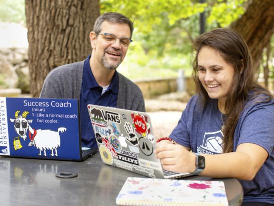 The image shows a man and a young woman sitting at an outdoor table, both using laptops. The man, wearing glasses and a cardigan, has a laptop with a sticker that reads "Success Coach: Like a normal coach but cooler" along with a goat mascot. The woman, wearing a blue t-shirt, has a laptop covered in various stickers. They are both smiling and appear to be in a positive and engaging discussion. The background features a tree and a natural outdoor setting, suggesting a relaxed, supportive atmosphere.