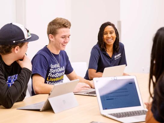 The image shows a group of four people sitting at a table, engaging in a discussion. They are all using laptops and appear to be in a modern, bright room. One person is wearing a baseball cap and a black shirt, another person is wearing a navy blue shirt with white text, a third person with braided hair is wearing a navy blue polo shirt, and the fourth person's face is not visible. They all seem to be in a positive and collaborative atmosphere, smiling and talking.