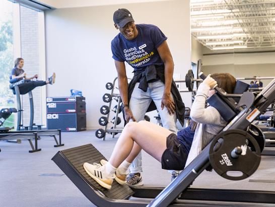 A peer training coach stands gives a student instructions on a hack squat press. Another student is in the background performing hanging leg raises.