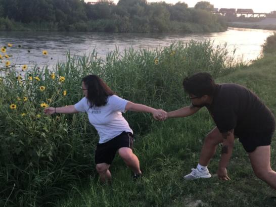 A man holding the hand of a woman to keep her from falling into a river as she reaches for a distant sunflower