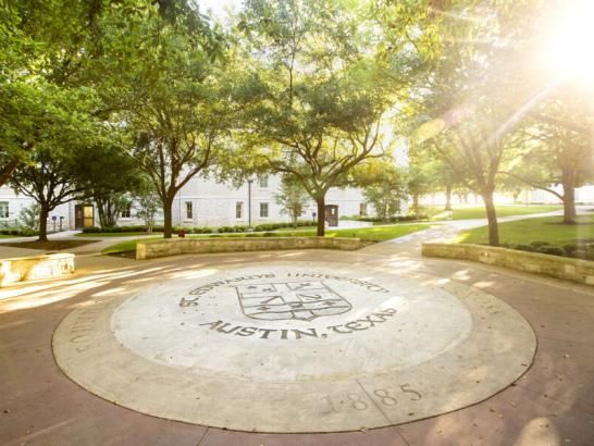 The image depicts a paved circular area with the words "ST. EDWARD'S UNIVERSITY AUSTIN, TEXAS" and the year "1885" inscribed on it. This emblem or seal is likely located on St. Edward's University campus in Austin, Texas. The surrounding lush green trees create a serene atmosphere, and white buildings with multiple windows can be seen in the background. The image conveys a sense of heritage and natural beauty associated with the university.