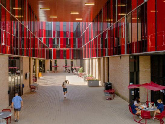 The image shows a vibrant courtyard area with striking red and black glass-paneled buildings. The space is framed by tall, modern structures creating a semi-enclosed atmosphere. People are walking and sitting at tables under red umbrellas, suggesting a casual and social environment. The ground is paved with bricks, and there are potted plants adding a touch of greenery. The design is contemporary and dynamic, with the red panels reflecting light, creating an engaging and lively atmosphere.