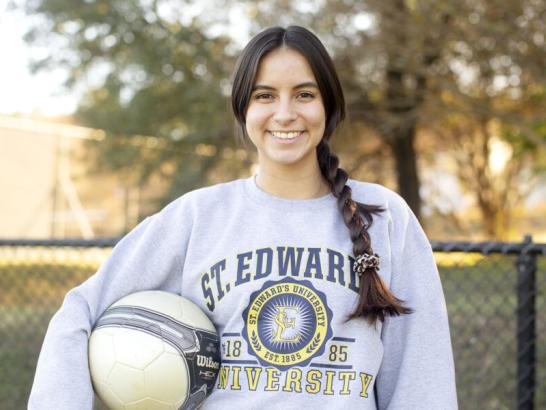 The image shows a young woman standing outdoors, holding a soccer ball under her right arm. She is smiling and wearing a grey sweatshirt with the text "St. Edward's University" and the university's logo on it. Her long hair is braided and tied with a scrunchie. The background features a fence and trees with autumn foliage, suggesting a sports or recreational setting at the university.