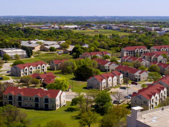 This image is an aerial view of a residential area featuring multiple buildings with red-tiled roofs and light-colored walls. The buildings are arranged in clusters surrounded by green lawns and trees, suggesting a suburban or campus setting. In the background, a broader view of the cityscape can be seen, with various buildings and infrastructure blending into the horizon under a clear blue sky. The scene conveys a sense of peacefulness and well-planned community living.