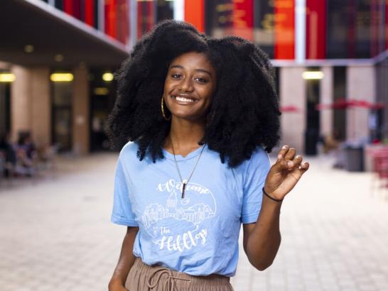 A student wearing a Welcome to the Hilltop shirt holds up a toppers up hand sign and stands in The Village patio area.