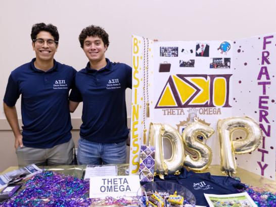 Two students stand behind a table featuring information for their business fraternity at the student involvement fair.