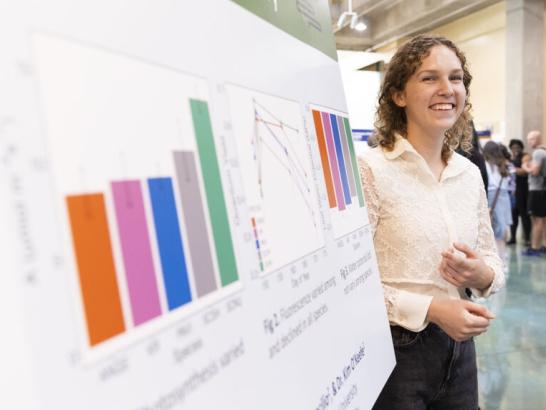 A student smiles as they stand next to their research posterboard depicting their findings with colorful bar graphs.