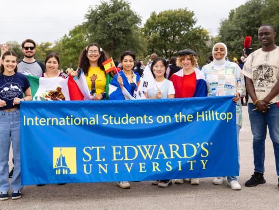 International Students stand in front of a banner that reads International Students on the Hilltop St. Edward's University before the homecoming parade.