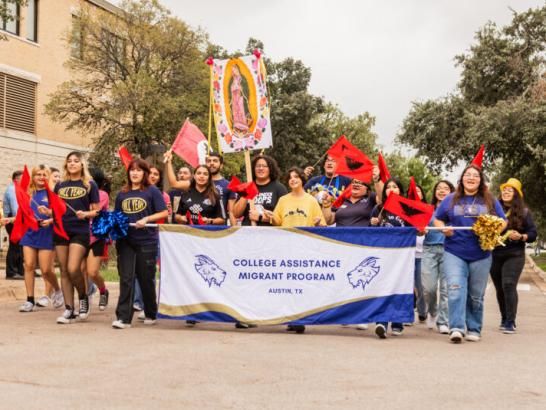 CAMP students and staff members march with flags and a blue and gold banner that says College Assistance Migrant Program Austin TX as part of the homecoming parade.