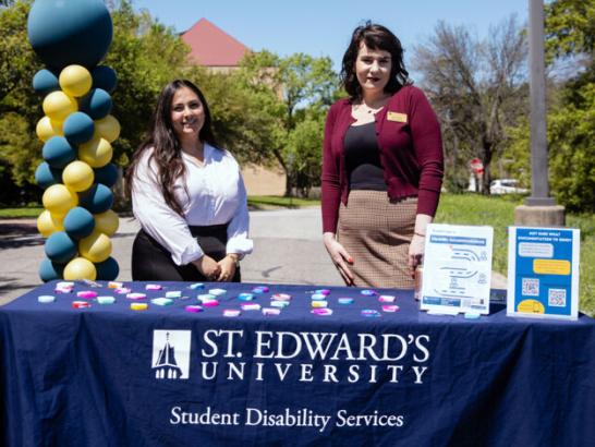 Two Student Disability Services staff members stand a table outside with informational posters and fidget toys. A blue and yellow balloon column and trees are in the background.