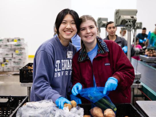 Two students hold produce and smile while volunteering at a food bank.