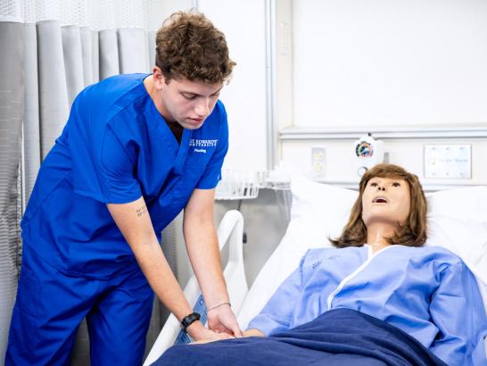 A student in scrubs takes the heart rate of a simulated manikin laying in a hospital bed