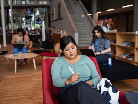 A student sits in a red chair writing on her tablet as other students study in the background at the Munday Library.