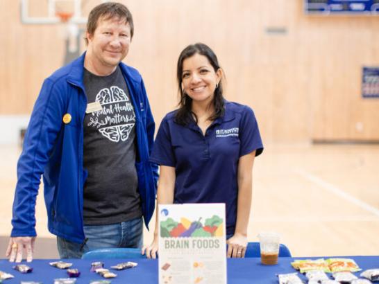 Health and Counseling Center staff members stand in front of a table that has a blue tablecloth and healthy snacks on it.