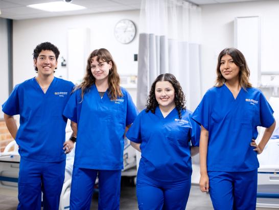 Four students wearing blue scrubs stand in a nursing simulation lab.