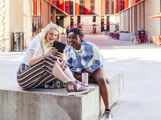 Two students sit outside in the Village patio area and smile as they look at a tablet.