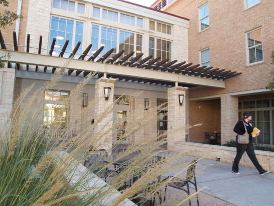 The image shows the exterior of a building with a mix of stone and brick construction. The entrance features large windows and a modern pergola, creating a welcoming and stylish facade. To the right, a person is walking while carrying books and a bag, suggesting a campus setting. In the foreground, ornamental grasses enhance the aesthetic appeal. There is outdoor seating with tables and chairs, indicating a comfortable space for socializing or studying. The environment is inviting and well-maintained.