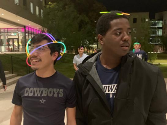 2 students walking across campus at night, wearing with glow sticks on their heads