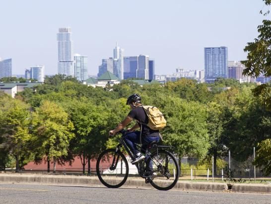 A student riding his bicycle on campus with downtown Austin skyline and greenery in the background. He wears a helmet, black t-shirt, jeans, sneakers and a backpack.